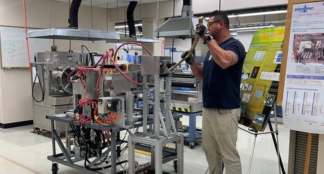 Physical Science Technician Wayne Swink feeds a hemp stalk into a lab-scale decorticating system, engineered by CQI staff at SRRC.  Electro-mechanical decorticators separate bast fibers from the woody core (hurd) of stalks.  Identifying the energy requirements of that process will help stakeholders in the hemp industry process raw hemp biomaterials more efficiently.